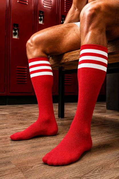 Red athletic knee-high socks with three white stripes near the top, worn on a person's legs while sitting on a wooden bench in a locker room with red lockers and wood flooring.